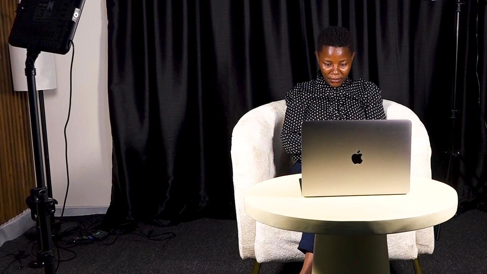 A presenter working at a laptop on the Kontext Media studio floor in Kampala, against a black curtain, lit by an LED panel