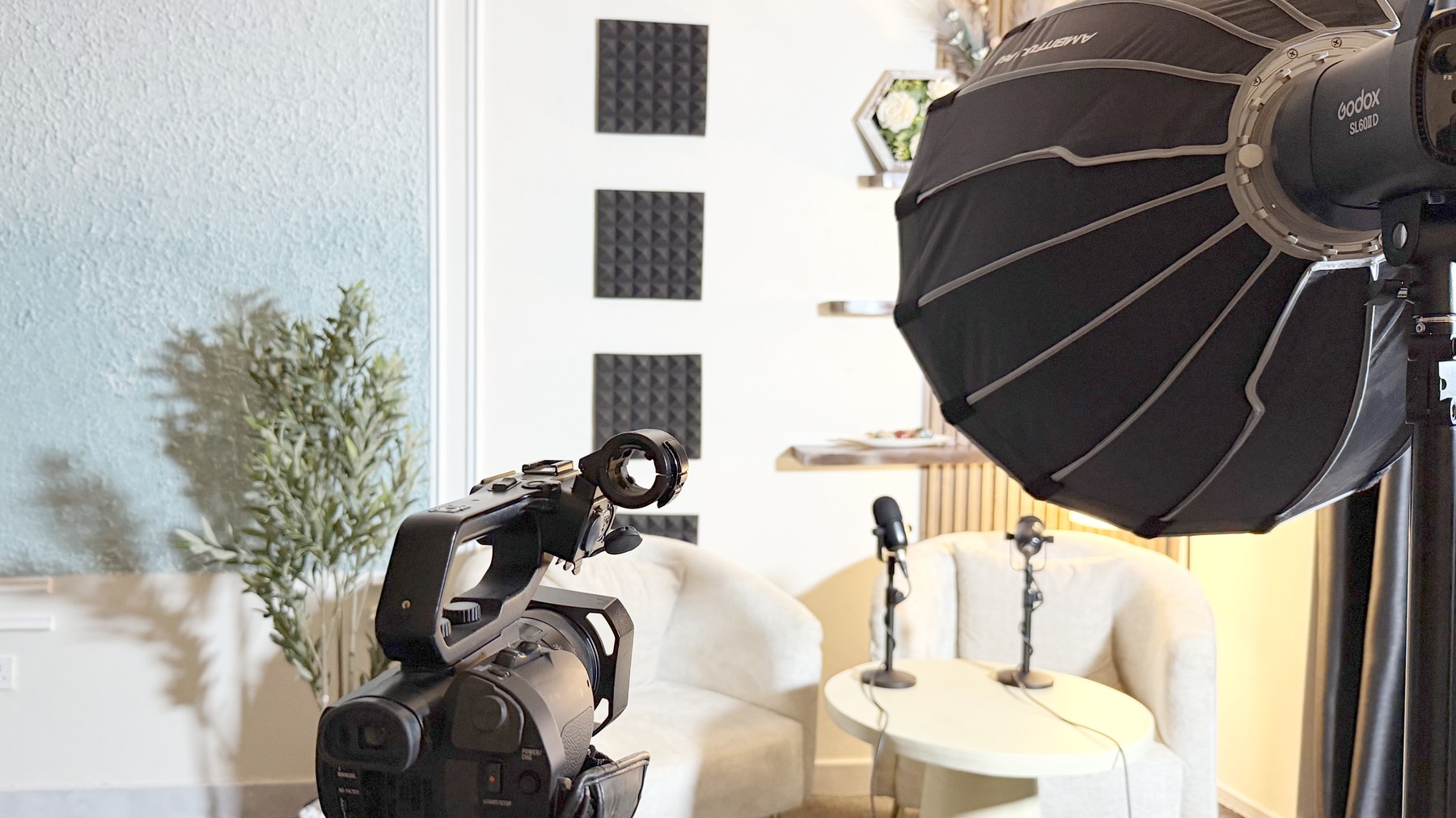 The Kontext Media studio in Kampala: two interview chairs and a low table with microphones, acoustic panelling and a slatted wood wall behind, lit by a Godox softbox, with a camera on a tripod in the foreground